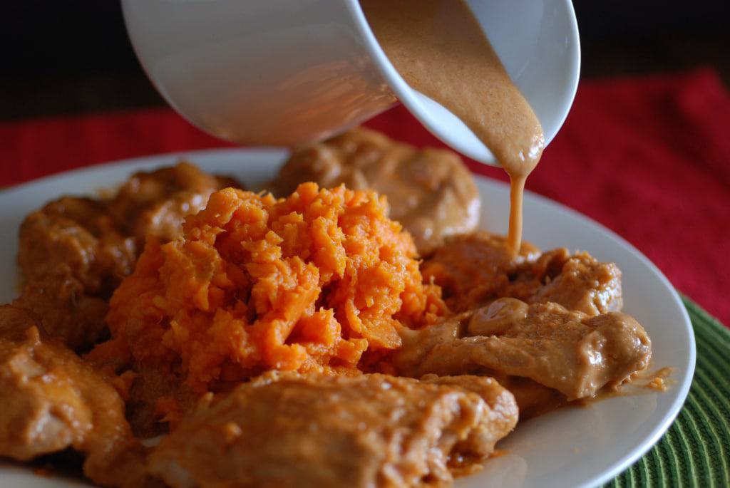 A picture of chicken thighs being drizzled with a creamy peanut sauce and served with mashed sweet potatoes.