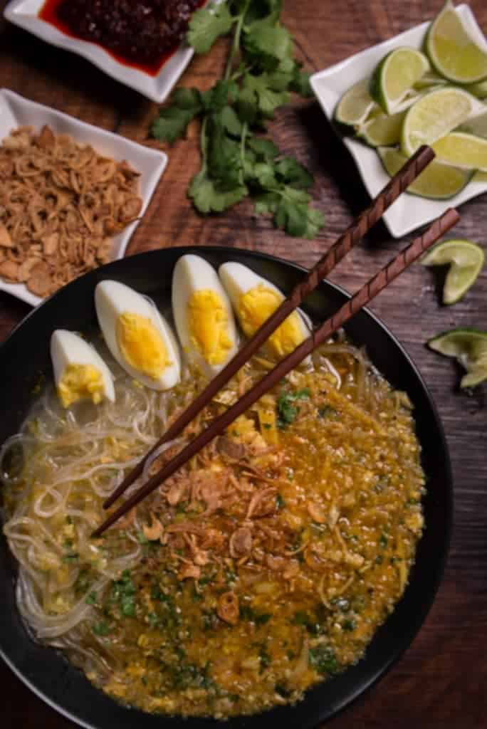 A picture of Myanmar Mohinga soup a warm fish broth and noodles with lots of condiments like lime, eggs, fried onions and chili paste
