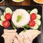 A plate of Hainanese chicken rice with three sauces, surrounded by cucumber and tomato slices.