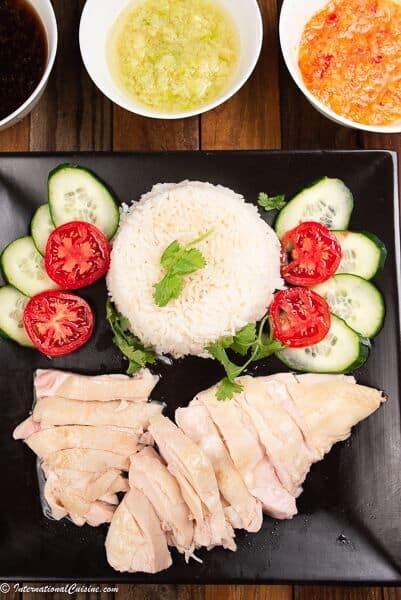 A plate of Hainanese chicken rice with three sauces, surrounded by cucumber and tomato slices.