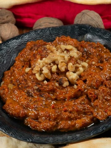 A bowl full of muhammara, a syrian red pepper sauce garnished with walnuts and surrounded by pita.