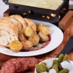 A raclette grill with melted bubbly cheese ready to put on boiled potatoes, bread, cured meat and gherkins with onions.