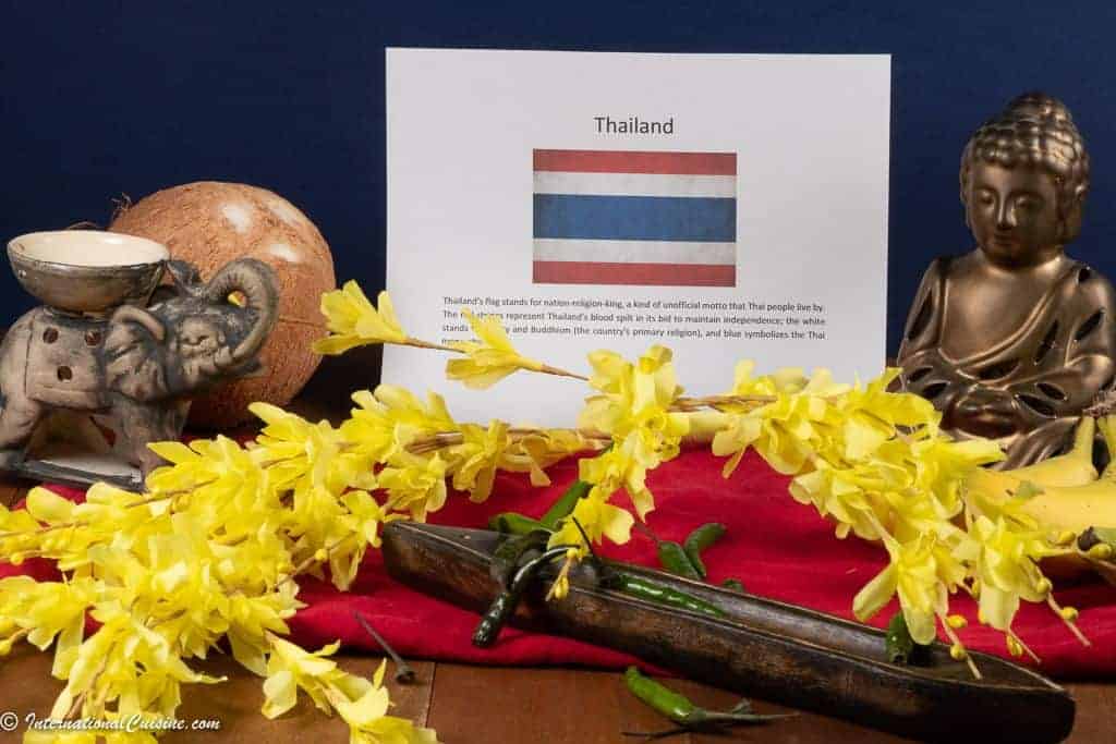 A menu with the Thai flag and symbols of Thailand a wooden boat, buddha statue, an elephant candle holder, a coconut and some Thai chilies.