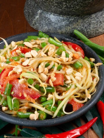 A bowl full of Thai green papaya salad with tomatoes, long green beans and Thai chilies.