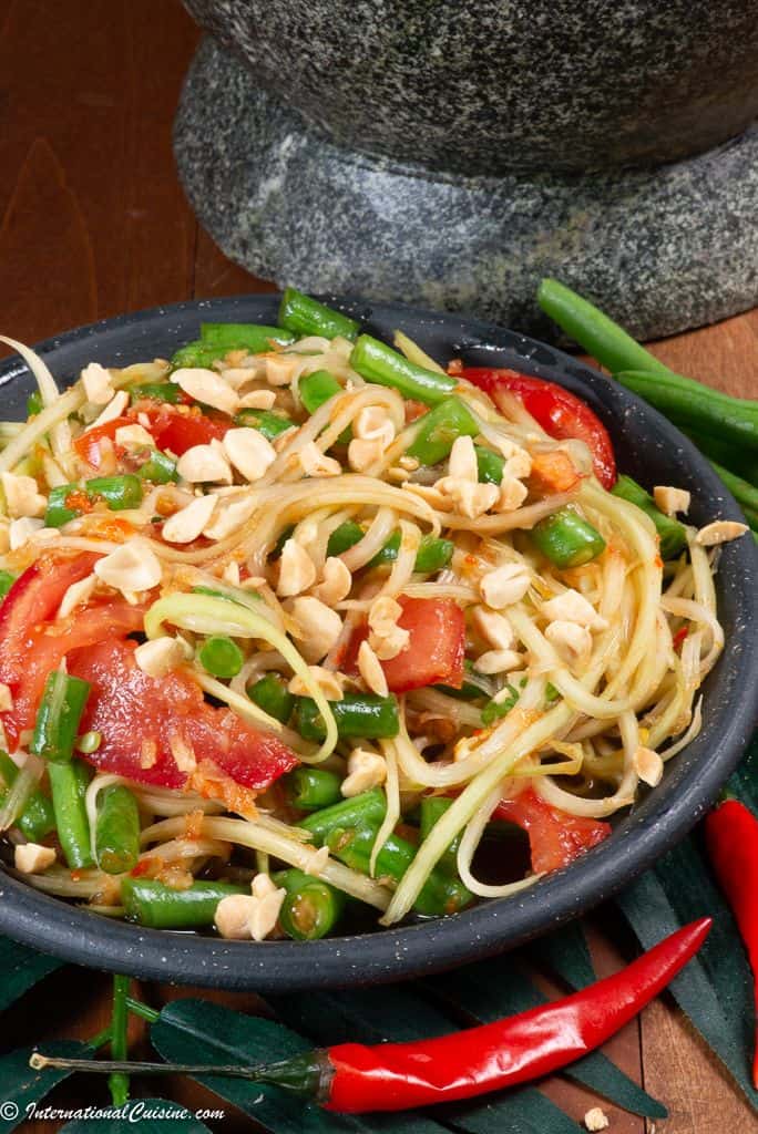 A bowl full of Thai green papaya salad with tomatoes, long green beans and Thai chilies.