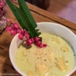 a bowl of Thai bananas in coconut milk sprinkled with sesame seeds and garnished with a pandan leaf and bright pink flowers.
