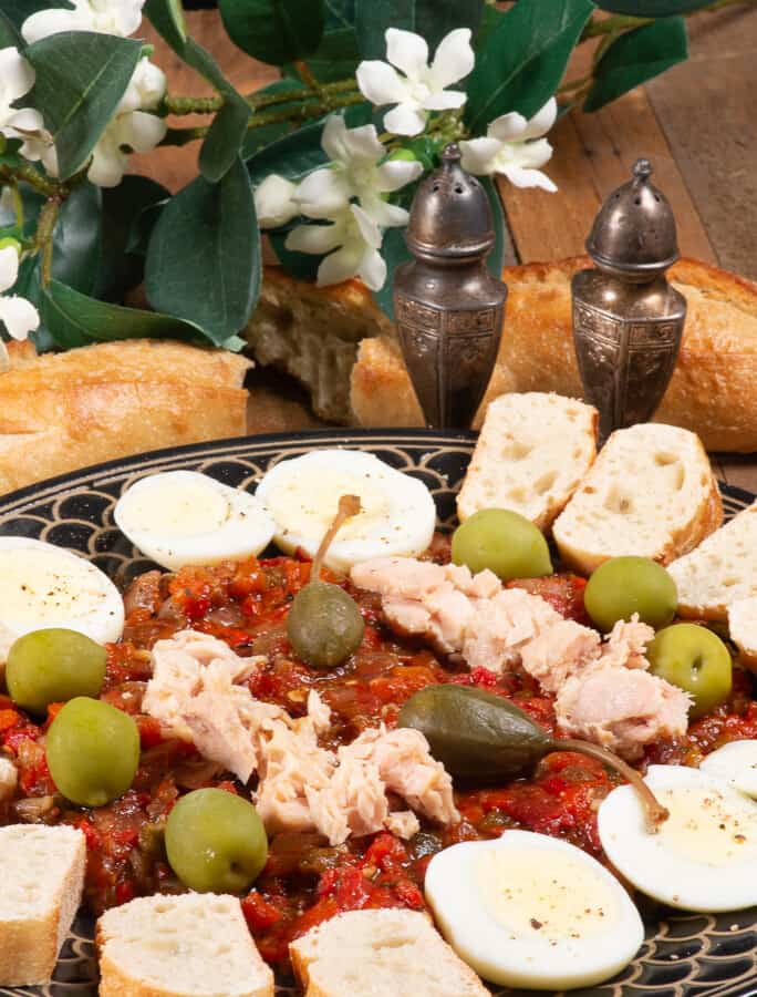 a plate of Mechouia salad topped with tuna and capers, surrounded by hard boiled eggs and French bread.